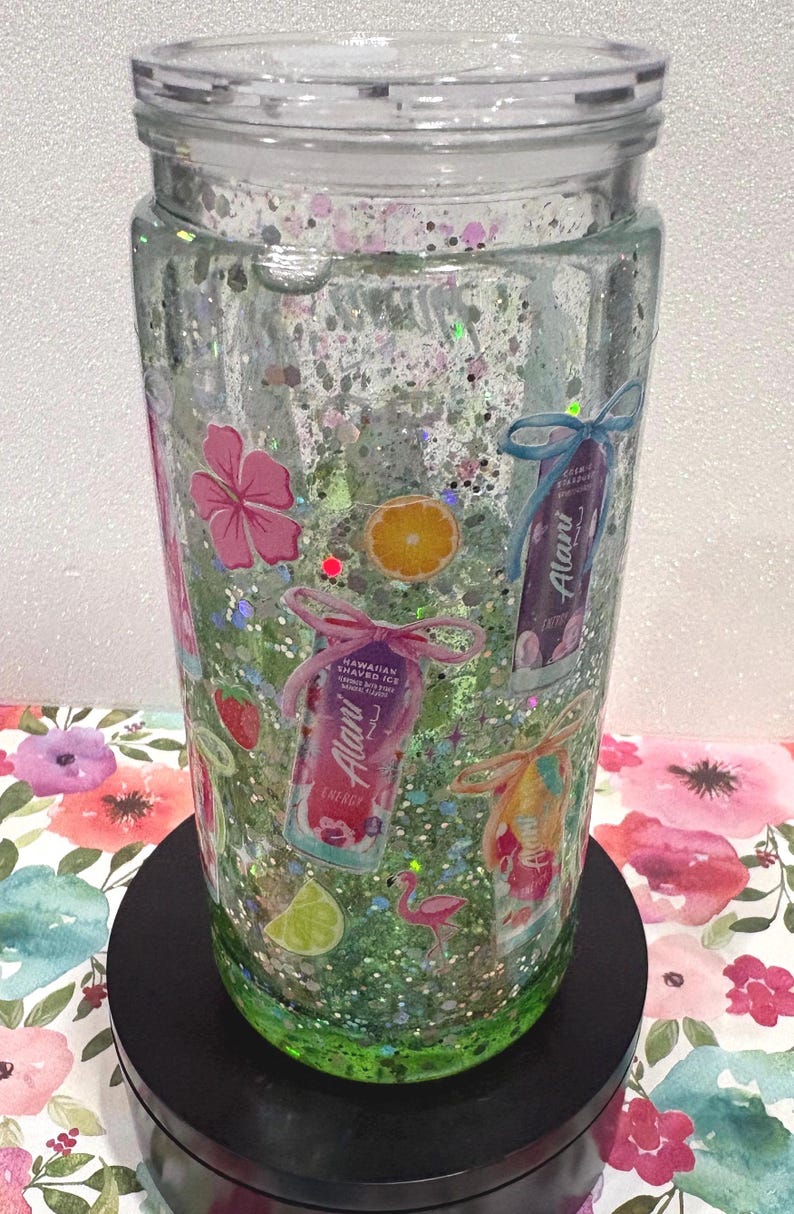 May include: A clear double-walled tumbler filled with green liquid and silver glitter. The exterior features decals of Alani energy drink cans, pink hibiscus flowers, orange and lime slices, strawberries, and a small pink flamingo graphic.