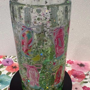 May include: A clear, double-walled tumbler filled with green liquid and iridescent glitter. The exterior features multiple small graphics of Alani energy drink cans, each tied with a decorative bow, along with small lime wedge illustrations.