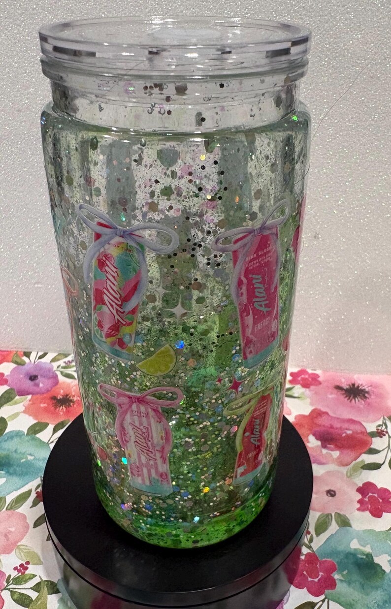 May include: A clear, double-walled tumbler filled with green liquid and iridescent glitter. The exterior features multiple small graphics of Alani energy drink cans, each tied with a decorative bow, along with small lime wedge illustrations.