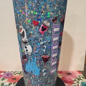 May include: A clear tumbler filled with blue liquid and iridescent glitter, featuring a graphic of Olaf, small snowgies, a castle, and various treats. The side includes a printed checklist with labels for Decaf, Shots, Syrup, Milk, Custom, and Drink.