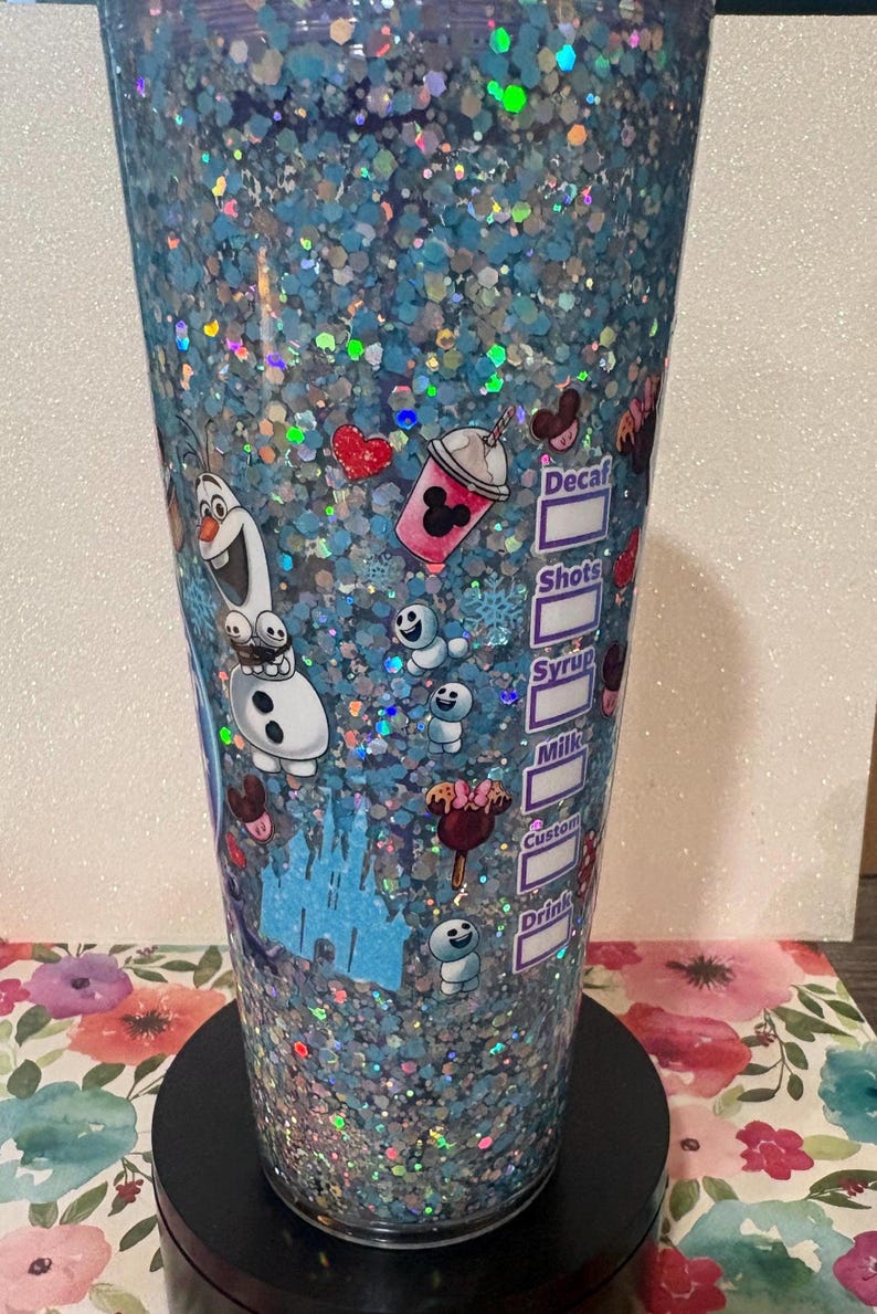 May include: A clear tumbler filled with blue liquid and iridescent glitter, featuring a graphic of Olaf, small snowgies, a castle, and various treats. The side includes a printed checklist with labels for Decaf, Shots, Syrup, Milk, Custom, and Drink.