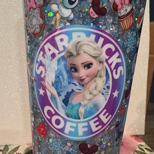May include: A cylindrical tumbler filled with light blue glitter and decorated with various character decals, including a central circular logo featuring a blonde character in a blue dress with the text STARBUCKS COFFEE surrounding the image.