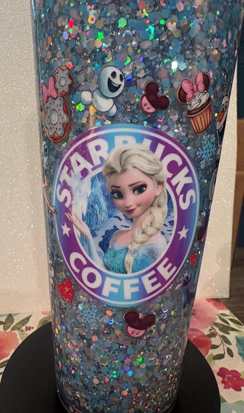 May include: A cylindrical tumbler filled with light blue glitter and decorated with various character decals, including a central circular logo featuring a blonde character in a blue dress with the text STARBUCKS COFFEE surrounding the image.