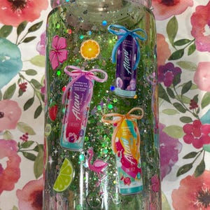 May include: A clear cylindrical tumbler filled with green liquid and iridescent glitter. The exterior features decals of three Alani Nu energy drink cans, a pink flamingo, a lime slice, an orange slice, a strawberry, and pink hibiscus flowers.