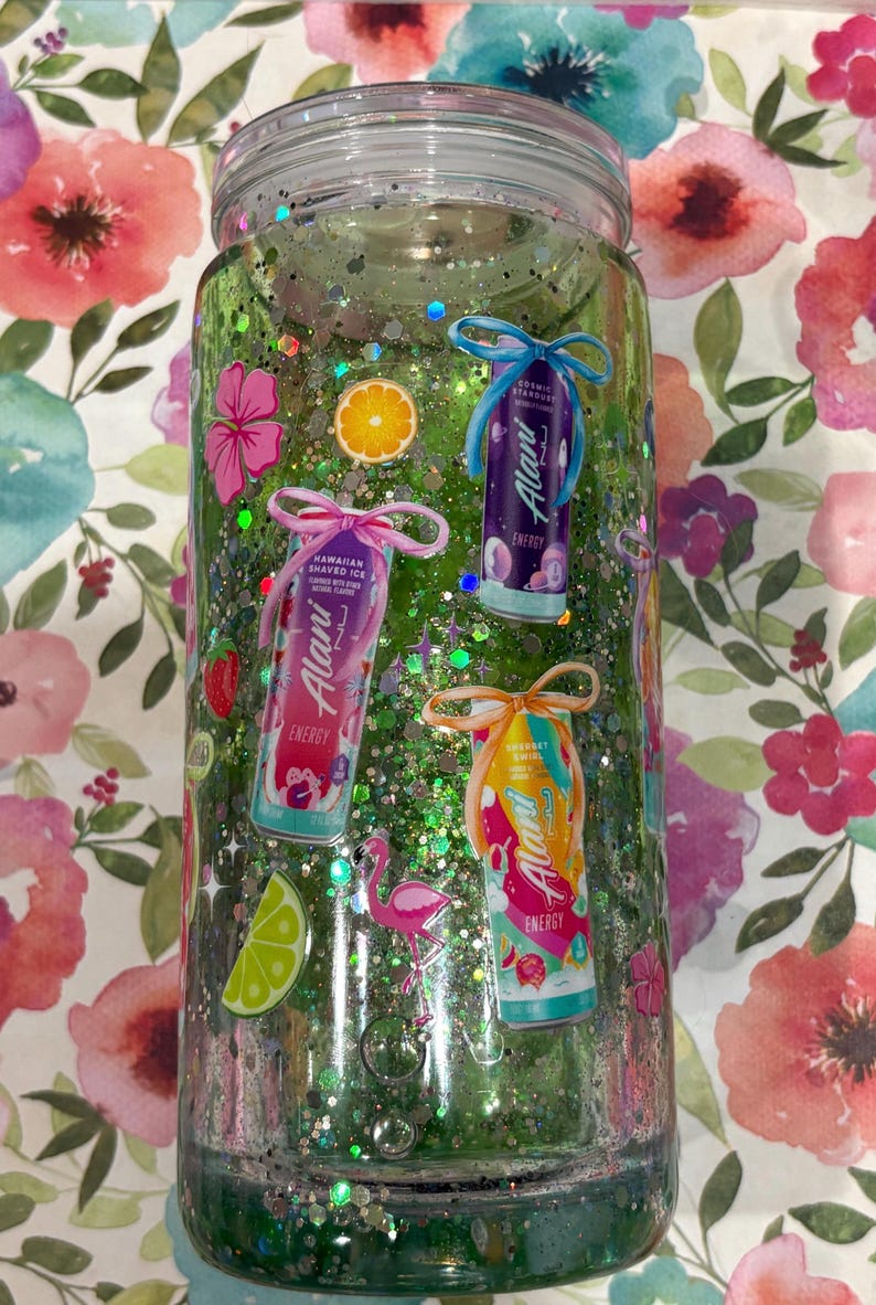 May include: A clear cylindrical tumbler filled with green liquid and iridescent glitter. The exterior features decals of three Alani Nu energy drink cans, a pink flamingo, a lime slice, an orange slice, a strawberry, and pink hibiscus flowers.