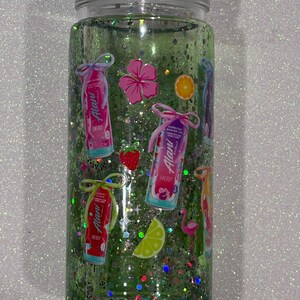 May include: A clear cylindrical tumbler filled with green liquid and iridescent glitter. The exterior features various stickers including Alani energy drink cans, a pink hibiscus flower, a strawberry, an orange slice, a lime wedge, and a pink flamingo.