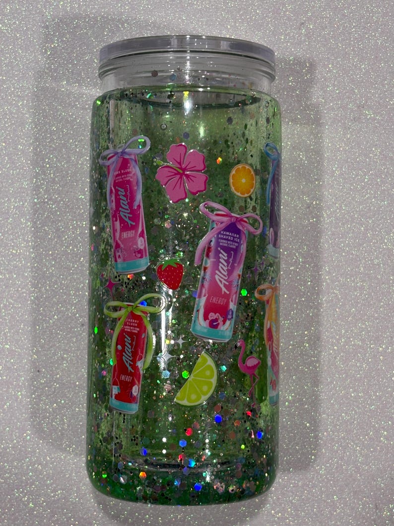 May include: A clear cylindrical tumbler filled with green liquid and iridescent glitter. The exterior features various stickers including Alani energy drink cans, a pink hibiscus flower, a strawberry, an orange slice, a lime wedge, and a pink flamingo.