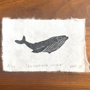 Puede incluir: Una impresi&oacute;n en lin&oacute;leo en blanco y negro de una ballena jorobada sobre un fondo de papel blanco. La impresi&oacute;n est&aacute; numerada 5/50 y firmada "HRF 25".
