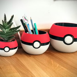 May include: Three red, white and black Pokeball-shaped planters. The smallest planter has a small green succulent plant inside. The middle planter has a collection of pens and pencils inside. The largest planter is empty.