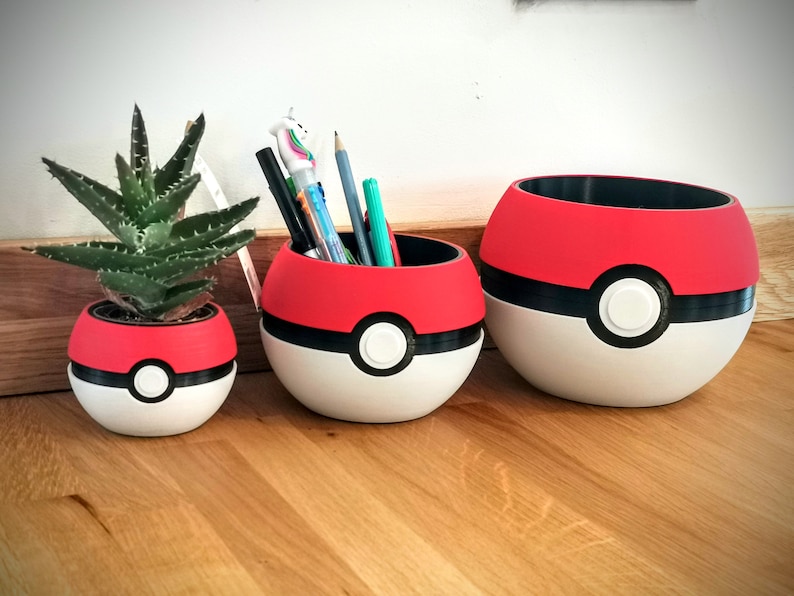 May include: Three red, white, and black Pokeball-shaped planters. The smallest planter has a small green succulent plant inside. The middle planter has a collection of pens and pencils inside. The largest planter is empty.