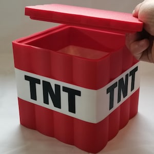 May include: Red plastic box with a white stripe and the word "TNT" on each side. The box has a lid that is open. The box is a replica of a Minecraft TNT block.