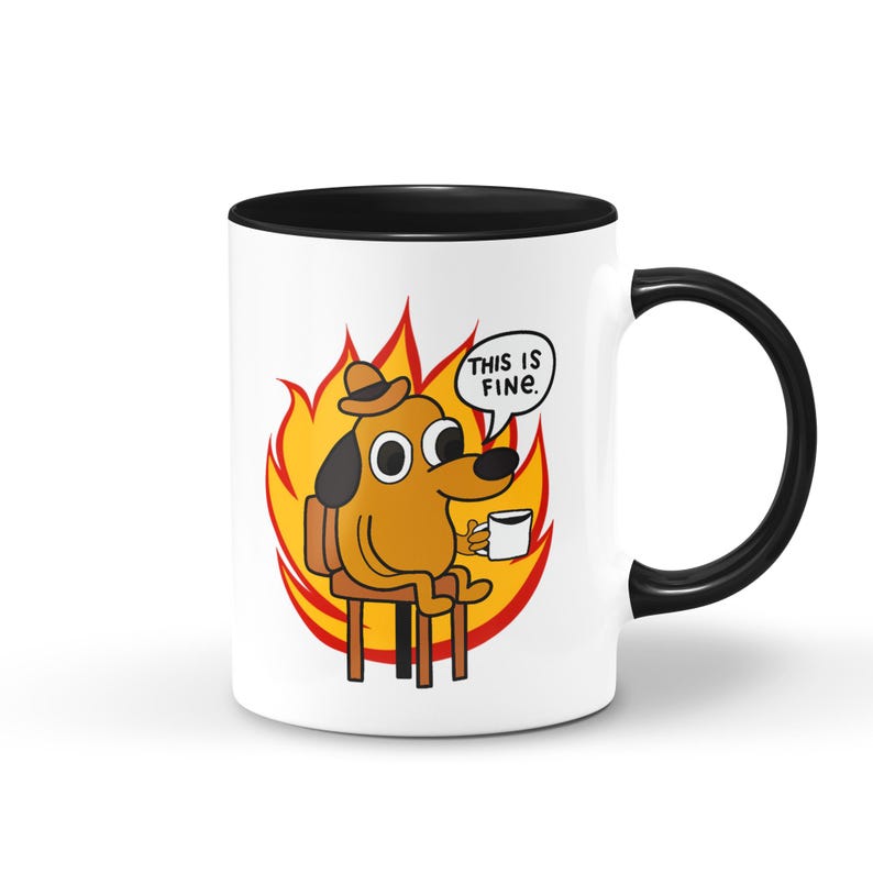 May include: White ceramic mug with a black rim and handle. The mug features a cartoon dog sitting in a chair surrounded by flames. The dog is wearing a hat and holding a cup of coffee. The text "This is fine." is written in a speech bubble above the dog.