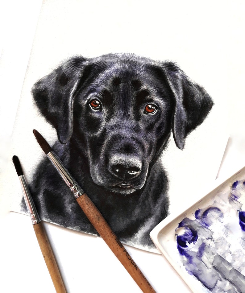 May include: Watercolor painting of a black Labrador Retriever puppy with brown eyes. The painting is on white paper and there are two paintbrushes and a paint palette in the background.