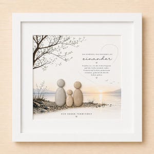 Könnte beinhalten: Ein gerahmtes Wandbild mit einer Kieselstein-Familie aus drei Figuren am Seeufer. Der Hintergrund zeigt einen Sonnenuntergang über dem Wasser mit einem Baumzweig. Der Text lautet: DAS SCHÖNSTE, WAS WIR HABEN, IST einander. FÜR IMMER VERBUNDEN.