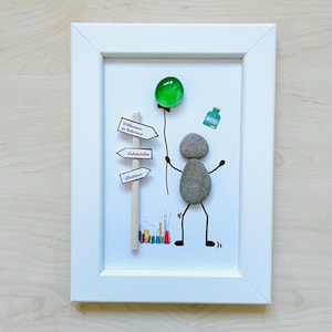 Könnte beinhalten: Gerahmtes Kunstwerk mit einer Strichmännchen-Figur aus Steinen, die einen grünen Glasballon hält. Die Figur steht in der Nähe eines Wegweisers mit deutschem Text und einer kleinen Flasche mit der Aufschrift "ACID".