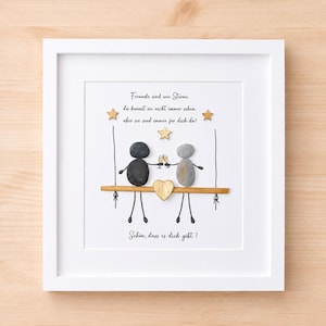 Könnte beinhalten: Ein weiß gerahmtes Wandbild mit Kieselsteinkunst von zwei Figuren, die auf einer Schaukel sitzen und Sektgläser halten. Das Design enthält drei goldene Sterne, ein goldenes Herz und den Text: "Freunde sind wie Sterne, du kannst sie nicht immer sehen, aber sie sind immer für dich da! Schön, dass es dich gibt!"
