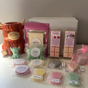 May include: A collection of wax melts in various shapes and colors, including heart and highland cow designs, alongside a ceramic highland cow wax warmer. The set features branded packaging with labels like Sunday Cuddles, Clean Queen, and Rhubarb Apple.