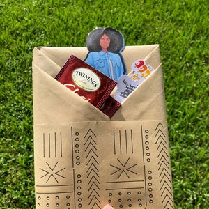 May include: A brown paper gift bag with a black and white geometric pattern. The bag is filled with a tea bag, a sticker, and a bookmark featuring a cartoon woman with an afro.