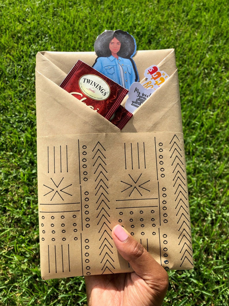 May include: A brown paper gift bag with a black and white geometric pattern. The bag is filled with a tea bag, a sticker, and a bookmark featuring a cartoon woman with an afro.