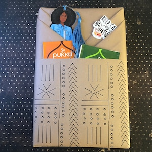 May include: A brown paper gift bag with a black and white geometric pattern. The bag is filled with a blue tassel bookmark with a black woman's illustration, a small orange box with the word "pukka" on it, and a small green tea bag with the text "Keep Calm and Be Sunshine".