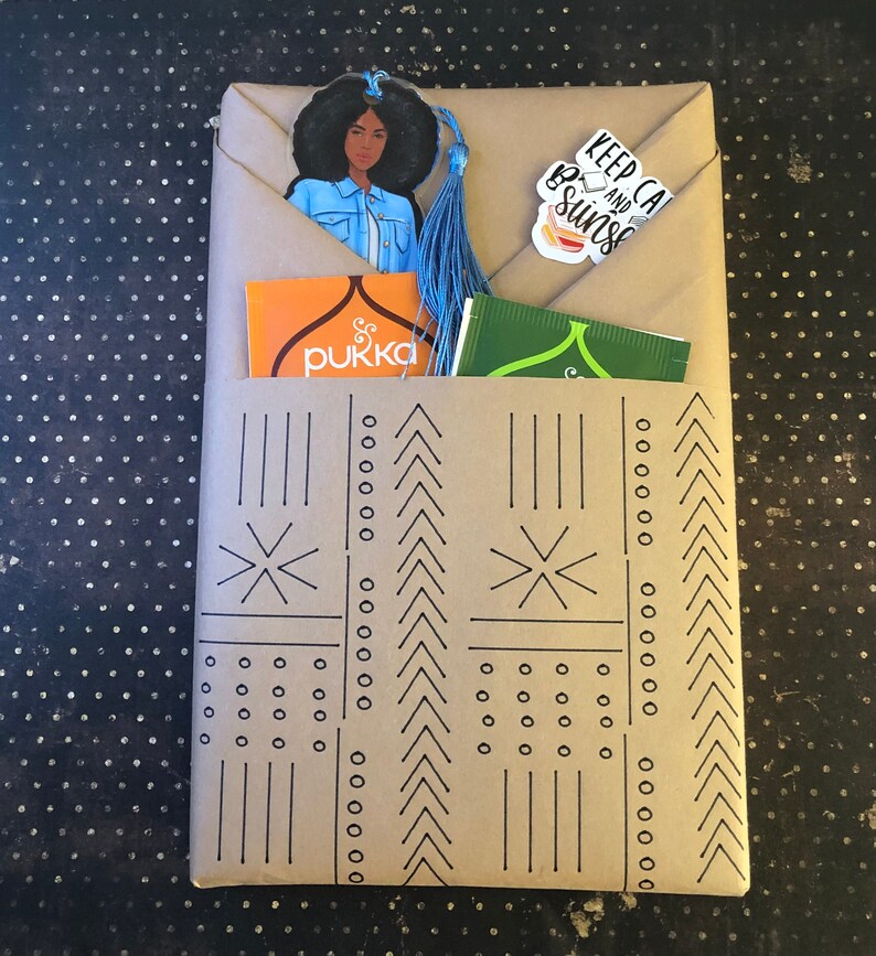 May include: A brown paper gift bag with a black and white geometric pattern. The bag is filled with a blue tassel bookmark with a black woman's illustration, a small orange box with the word "pukka" on it, and a small green tea bag with the text "Keep Calm and Be Sunshine".