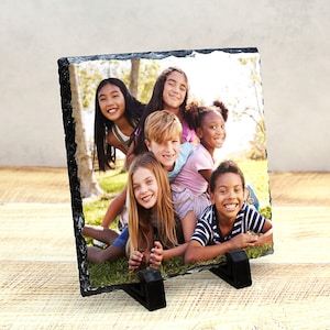 May include: A group of children smiling and posing for a photo. The children are standing in a grassy area with trees in the background. The photo is printed on a slate coaster with a rough, natural edge.