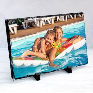 May include: Two children, a boy and a girl, are smiling and having fun on a white inflatable raft in a swimming pool. The pool is surrounded by a concrete deck with lounge chairs.