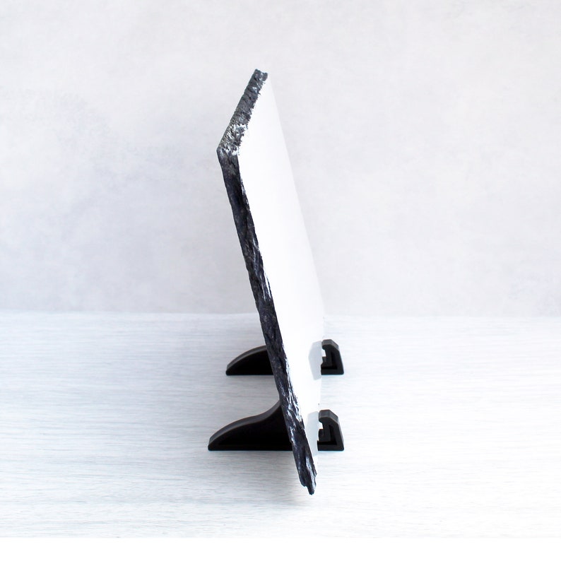 May include: A black slate photo frame with a white surface and a black stand. The frame is standing on a white surface.
