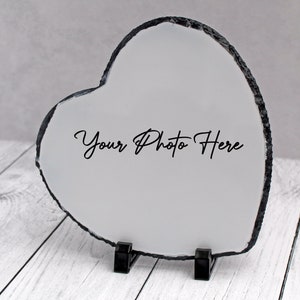May include: A white heart-shaped slate photo frame with a black border and the text "Your Photo Here" printed on it. The frame is standing on a white wooden surface.