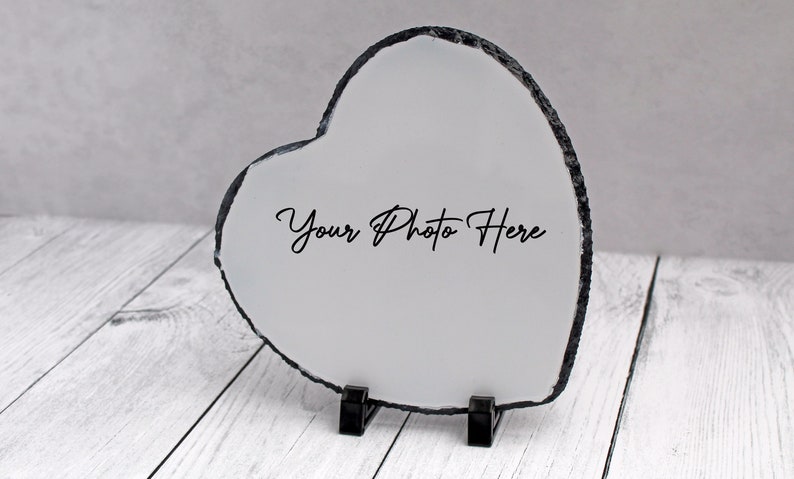 May include: A white heart-shaped slate photo frame with a black border and the text "Your Photo Here" printed on it. The frame is standing on a white wooden surface.