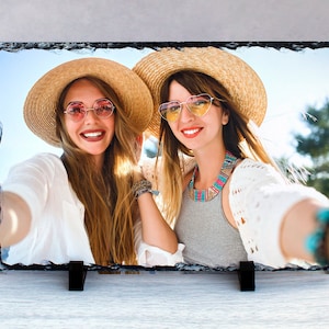 May include: Two women wearing straw hats and sunglasses are smiling and taking a selfie. They are both wearing white shirts and one is wearing a gray shirt with a colorful necklace.
