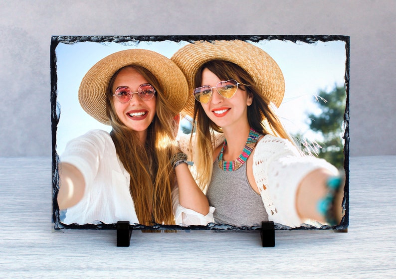 May include: Two women wearing straw hats and sunglasses are smiling and taking a selfie. They are both wearing white shirts and one is wearing a gray shirt with a colorful necklace.