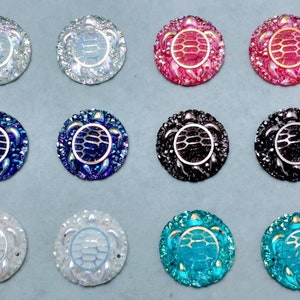 Peut inclure: Douze cabochons ronds en résine décoratifs en forme de tortue, dans différentes couleurs, dont le blanc, le bleu, le rose, le noir et le turquoise. Chaque cabochon présente un motif de tortue détaillé avec une carapace texturée.