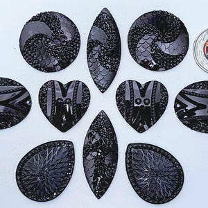 May include: A collection of black decorative embellishments in diverse shapes, such as circles, ovals, teardrops, and hearts. The embellishments feature detailed patterns and textures. A coin with a red and black design is also included.