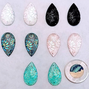 Peut inclure: Huit perles de verre en forme de larme avec des motifs floraux dans différentes couleurs, notamment le blanc, le noir, le bleu, le vert et le rose. Les perles sont disposées en deux rangées de quatre, avec une pièce canadienne de deux dollars en bas à droite pour l'échelle.