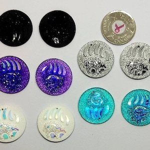 May include: Assortment of ten round bear paw print embellishments. The embellishments are in various colours including black, silver, purple, white, and turquoise. The embellishments are iridescent and have a glitter finish.