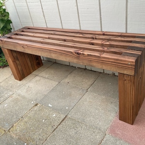 May include: A dark brown wooden bench with a slatted top and a simple design. The bench is made of rough-hewn wood and has a rustic look.