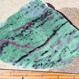 May include: A rough-cut stone with a vibrant green base, accented by streaks of black and red. A wooden ruler with inch markings is placed next to the stone. The stone's irregular shape and color variations create a unique, natural aesthetic.