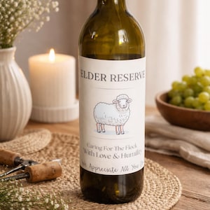 May include: A green wine bottle with a label that reads "ELDER RESERVE" and features a cartoon sheep. The label also includes the text "Caring For The Flock With Love & Humility We Appreciate All You Do". The bottle is on a wooden surface.