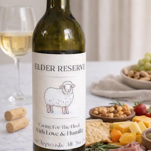 May include: A green wine bottle with a black cap and a label that reads "ELDER RESERVE." The label features a cartoon sheep and the text "Caring For The Flock With Love & Humility." A wine glass, corks, and a charcuterie board with snacks are in the background.