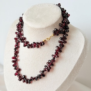 Op de afbeelding: Een ketting bestaande uit talloze kleine, dieprode, druppelvormige granaatkralen. De ketting is voorzien van een goudkleurige metalen karabijnsluiting die de twee uiteinden van de streng met elkaar verbindt tot een doorlopende cirkelvormige lus.