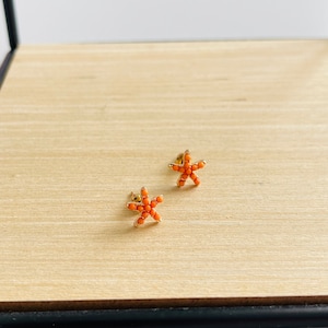 Könnte beinhalten: Ein Paar goldene Seestern-Ohrringe mit orangefarbenen Perlen. Die Ohrringe liegen auf einer Holzoberfläche.