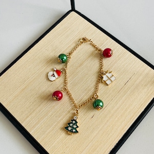 May include: Gold-toned charm bracelet featuring Christmas-themed charms. Charms include a Santa Claus, a Christmas tree, a gift box, and red and green baubles. The bracelet is displayed on a light wood surface.