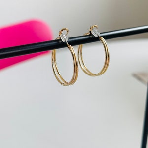 Könnte beinhalten: Ein Paar goldfarbene Creolen. Jeder Ohrring hat ein Doppelreifen-Design mit Clipverschluss. Die Ohrringe werden vor einem weißen Hintergrund mit einer schwarzen Stange präsentiert.