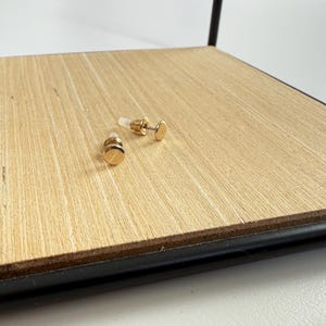 Könnte beinhalten: Zwei goldfarbene Ohrstecker mit flachem, kreisförmigem Design. Die Ohrringe werden auf einer hellen Holzoberfläche präsentiert. Die Ohrringe haben einen Stecker und einen Verschluss. Schmuck.