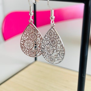 Könnte beinhalten: Silberfarbene, tropfenförmige Ohrringe mit floralem Filigranmuster. Die Ohrringe hängen an silbernen Haken und werden vor einem verschwommenen Hintergrund präsentiert. Ein zartes Accessoire.