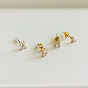 Könnte beinhalten: Drei goldene Ohrringe mit kleinen, klaren Strasssteinen in Form der Buchstaben K, D und L.