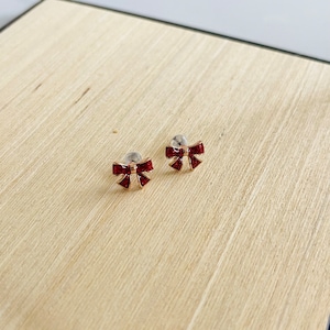 Puede incluir: Un par de pendientes de color dorado con dijes en forma de lazo rojo. Los lazos están adornados con piedras rojas brillantes.