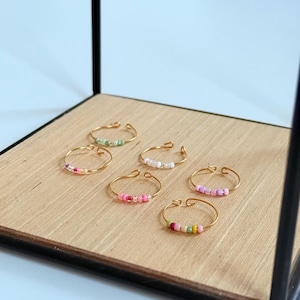 Könnte beinhalten: Sechs zarte goldfarbene Ringe, jeder mit einer Reihe farbiger Perlen verziert. Die Perlen sind in den Farbtönen Rosa, Grün, Weiß und Gelb gehalten. Die Ringe werden auf einer hellen Holzoberfläche auf einem schwarzen Metallständer präsentiert.