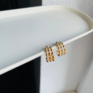 Könnte beinhalten: Ein Paar goldene Creolen mit einem strukturierten, perligen Design. Die Ohrringe sind klein und zart.